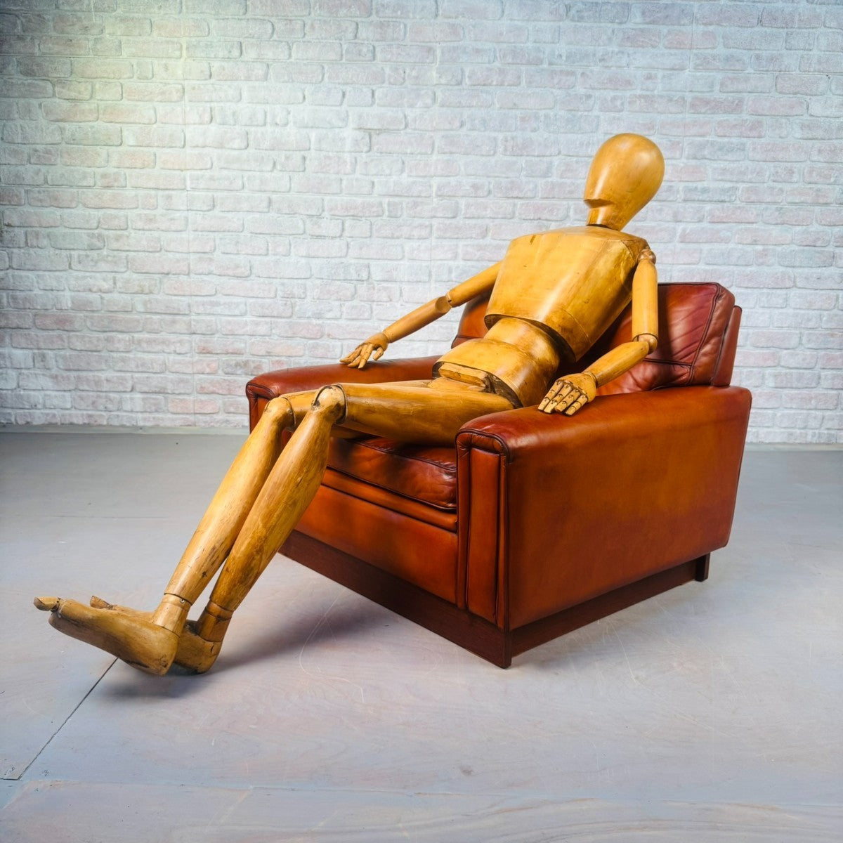 Wooden mannequin sitting on a brown leather armchair against a brick wall.